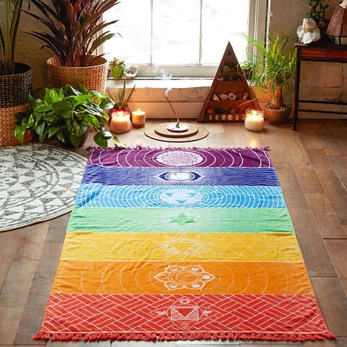A beautiful Yoga room with a brightly coloured mat in the center representing the chakras, surrounded by leafy pot plants and candles on a wood floor.
