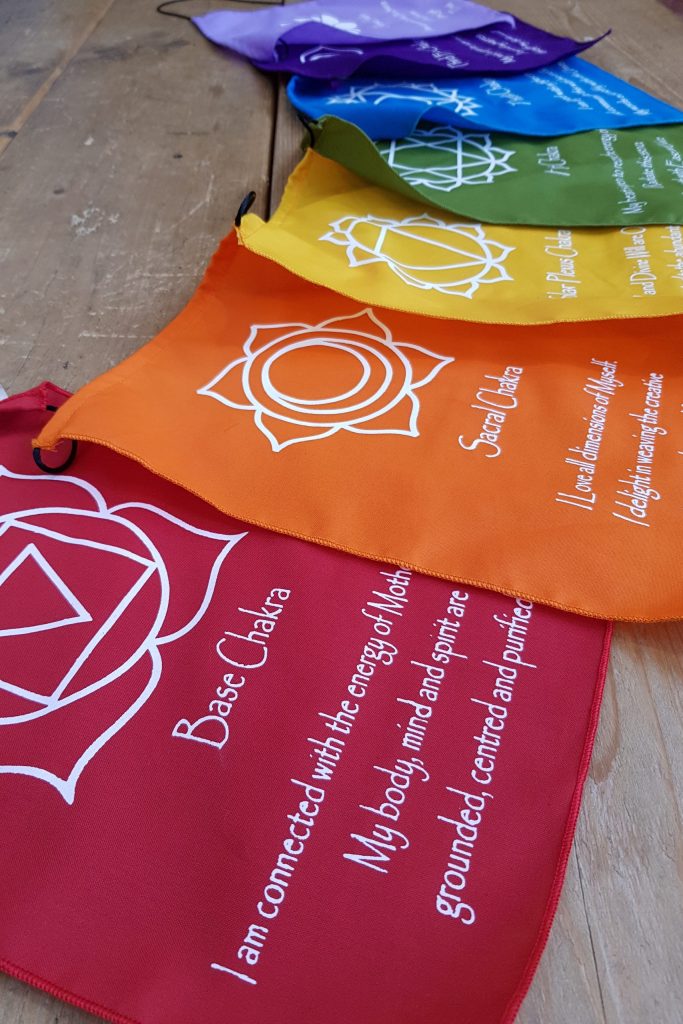 Colourful chakra flags laid out in a row each with a chakra symbol and affirmation in white printed on them
