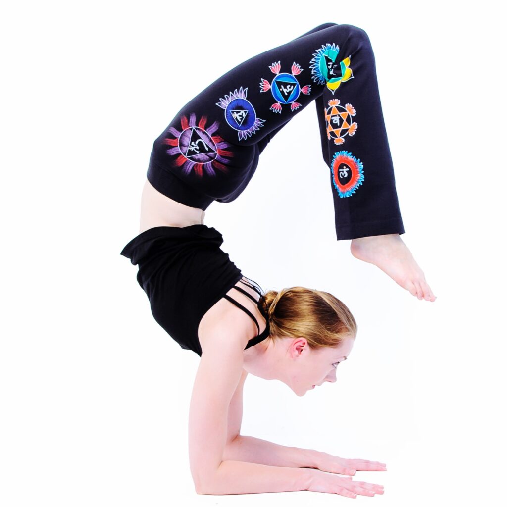 A yoga teacher in black yoga pants with painted chakras and a black camisole performing Scorpion Pose, an elbow stand with the spine curved and the feet over the head